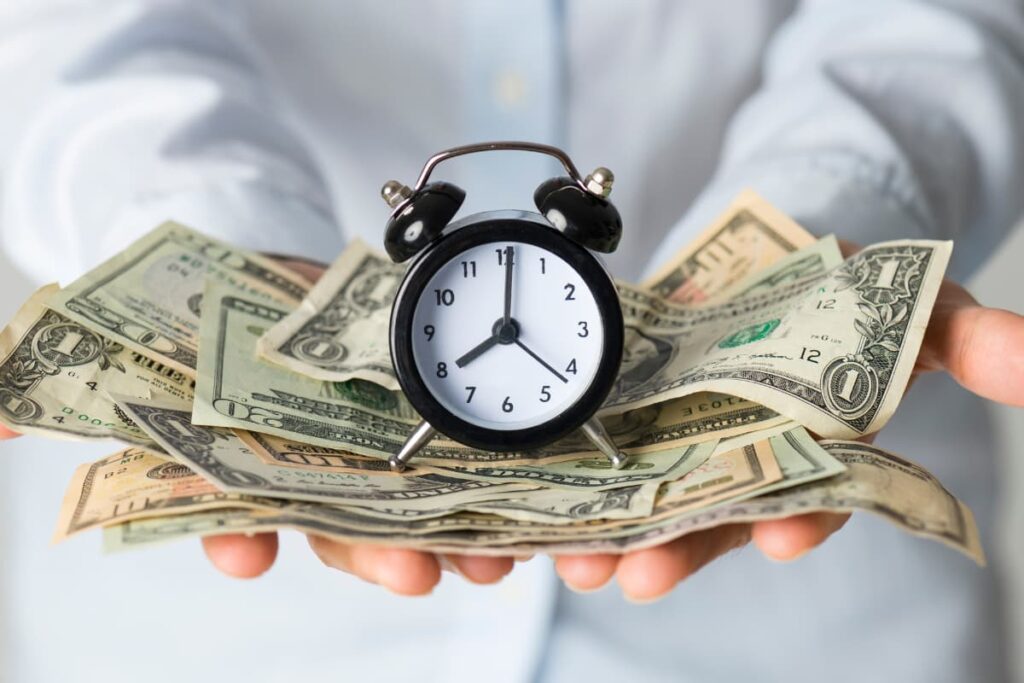 A Person wearing white shirt holds some dollars and a small Black color clock in his hands. Burger King opens at 8'0 AM in some locations.