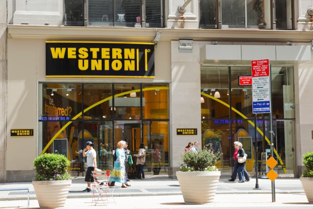 Around ten people are walking and two big size plant pots , one sitting chair and one direction board are in front of the Western Union block . 