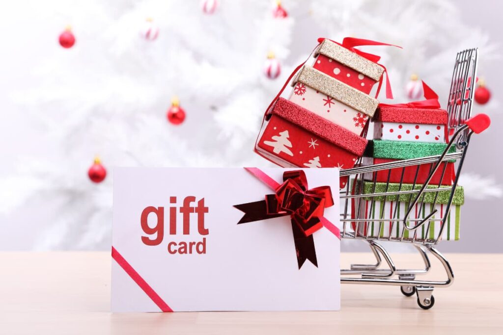 Image Consists of one gift card voucher, six decorative mini gift boxes in a small size cart with a backdrop of three red color Christmas decorative Ornaments.