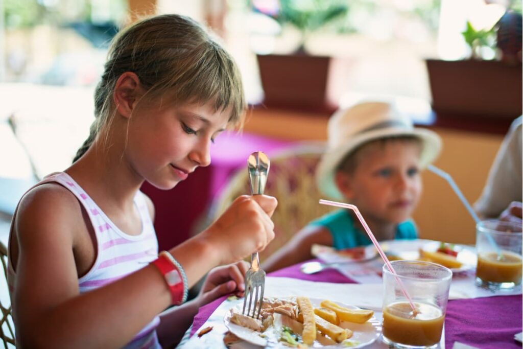 Wendy's Kids Meal is enjoying by 2 kids are having Wendy's Kids Meal which consists of a sandwich, few french fries in a white plate with fork and a chocolate drink in a glass having a straw in it.