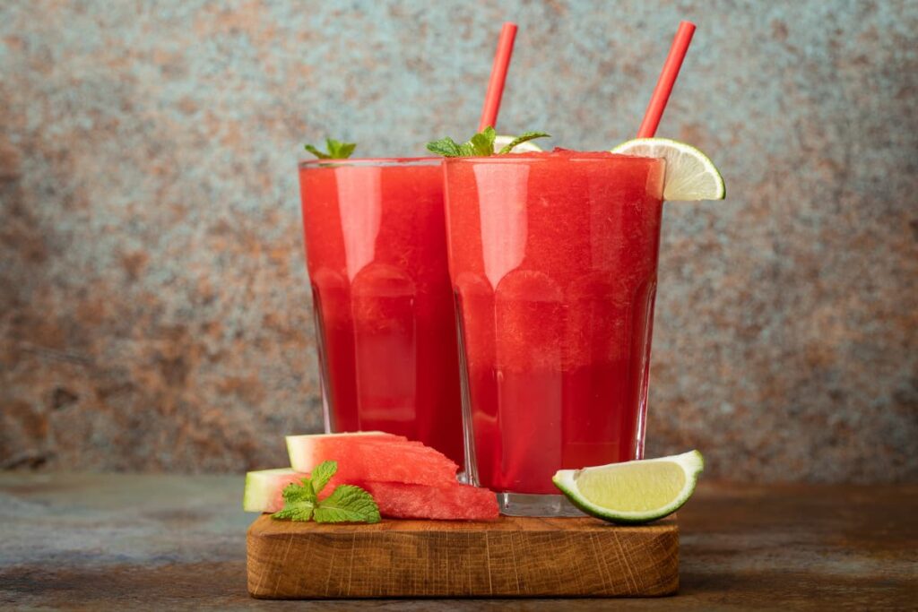 Wendy's Slushies has 2 glasses of Slushie with a slice of lemon and a mint leaves and a straws in it and 2 pieces of watermelon , 1 slice of lemon  and mint leaves on a wooden board.