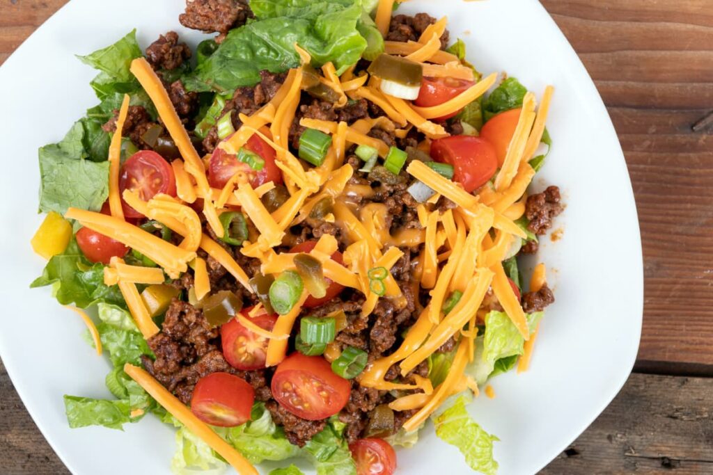 Wendy's Taco salad with has a Taco salad topping of few pieces of spring onions, few lettuce leaves, bunch of orange capsicum and few slices of tomato's served in a white plate on a table.