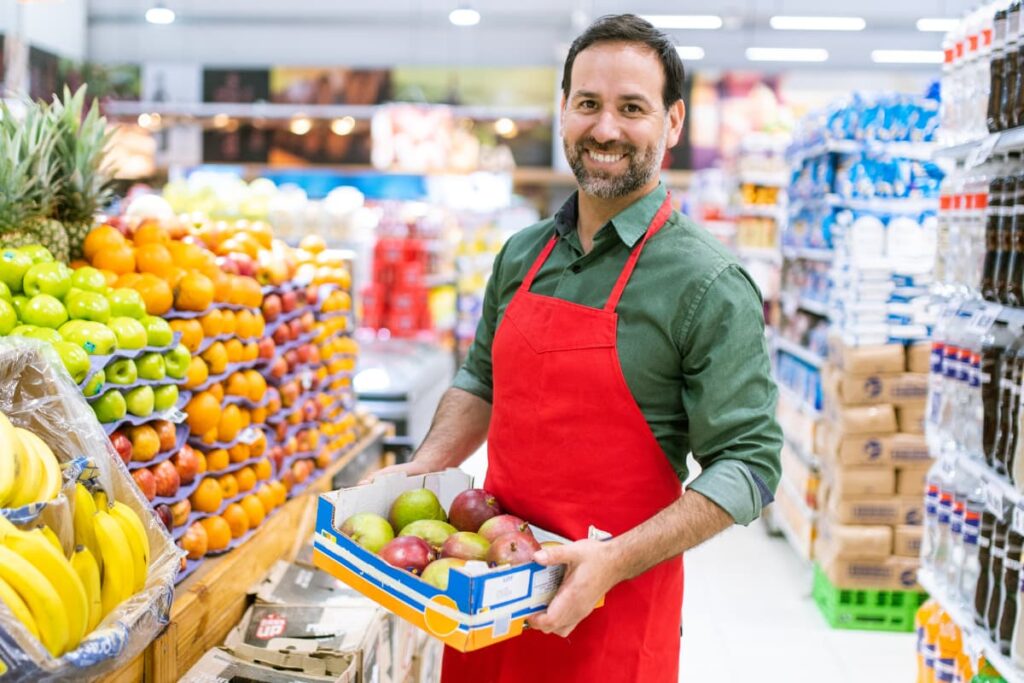 A Walmart Stocker wearing Red color apron and holding with a bunch of apples box at store and the stroe also have some bananas, oranges, green apples, water bottles. 