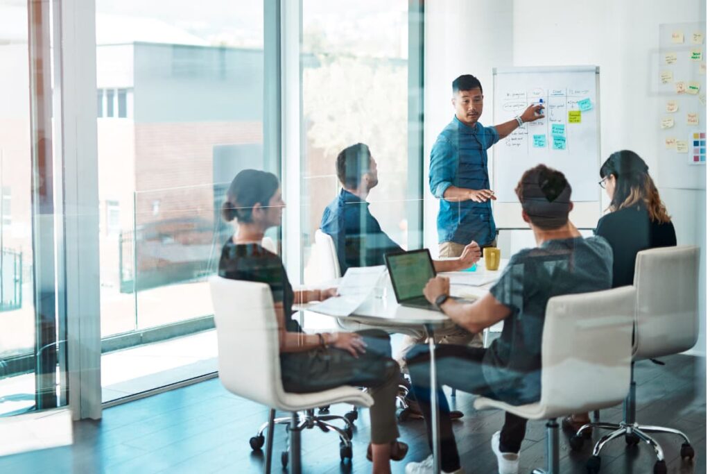 In a room consists of  employees, One member is discuss about business models in the white board and four members are sitting in the white chairs along with one laptop during Walmart Orientation program.