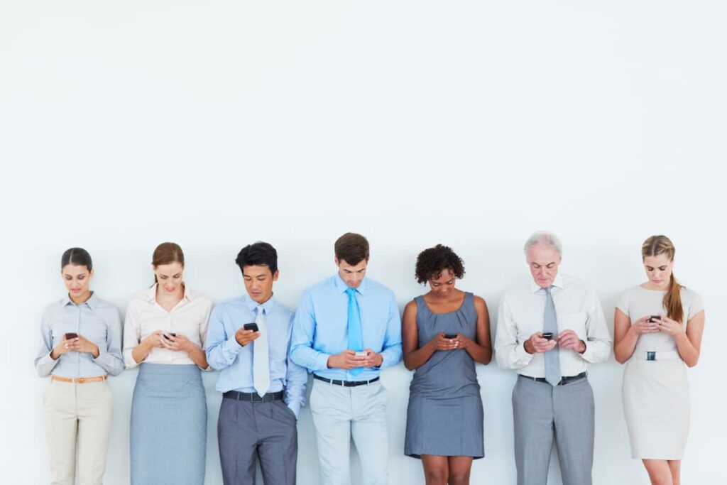 7 people are standing in a row, all are holding their mobiles. They all are above 18 years or older, all are different age group, wearing different style of dresses.
