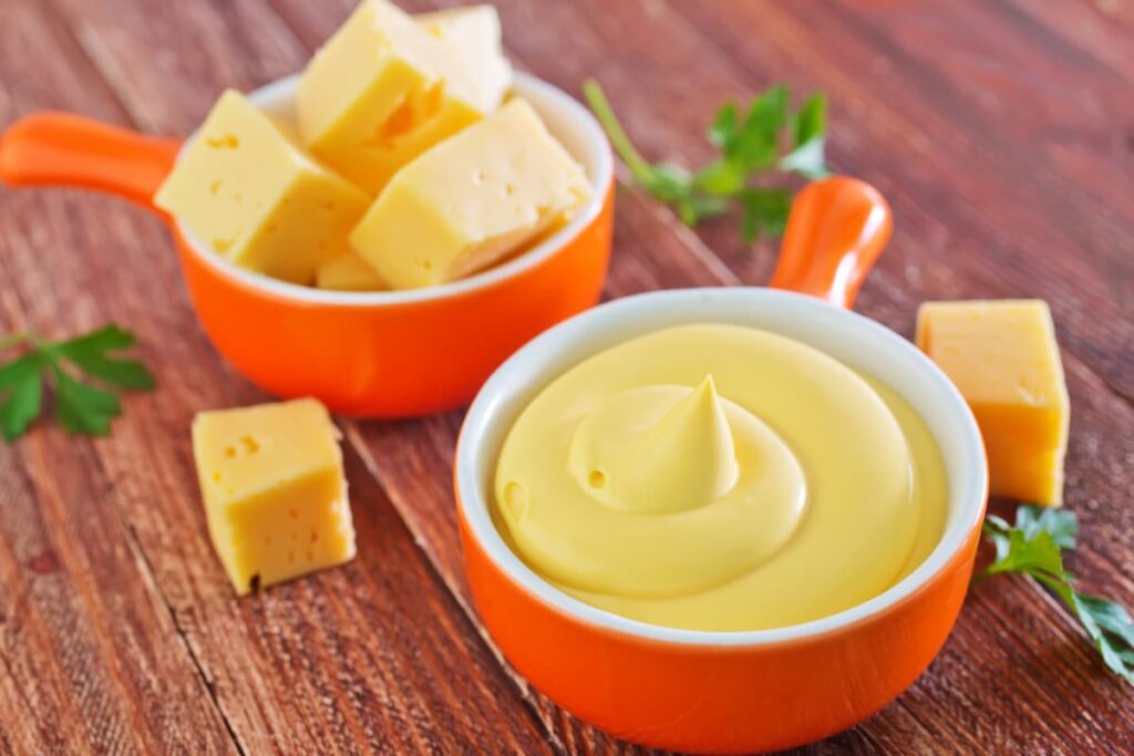Chick Fil A Cheese Sauce serves in a small orange color bowl. Besides there is another bowl which is having cheese cubes, 2 cheese cubes, and coriander leaves on the floor.