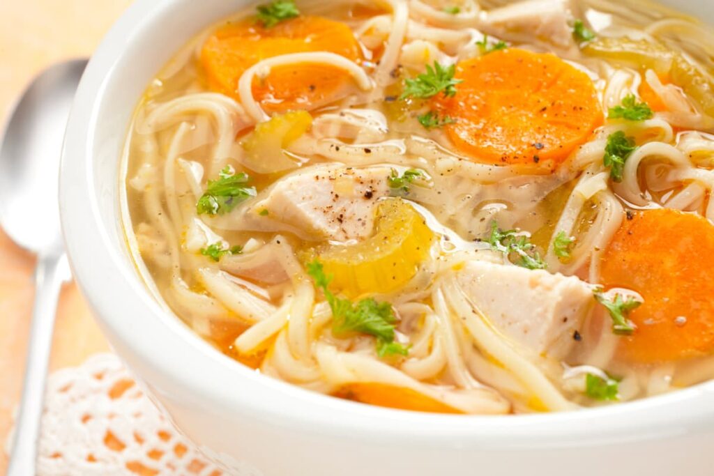 Chick Fil A Chicken Noodle Soup served in a white bowl along with white color spoon. This Chick Fil A Chicken soup contains boiled chicken, noodles, carrot slices, yellow capsicum slices, chicken broth and Coriander leaves for garnishing.