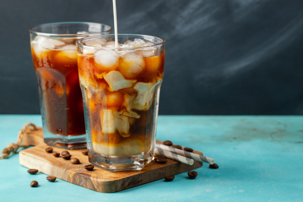 Chick Fil A Iced Coffee can be served in a brown color wooden plate. There are 2 glasses of iced coffee which has cream, red color syrup, caramel, coffee beans. Beside these 2 glasses of Chick fil a iced coffee there are some beans and 2 straws on the wooden plate.