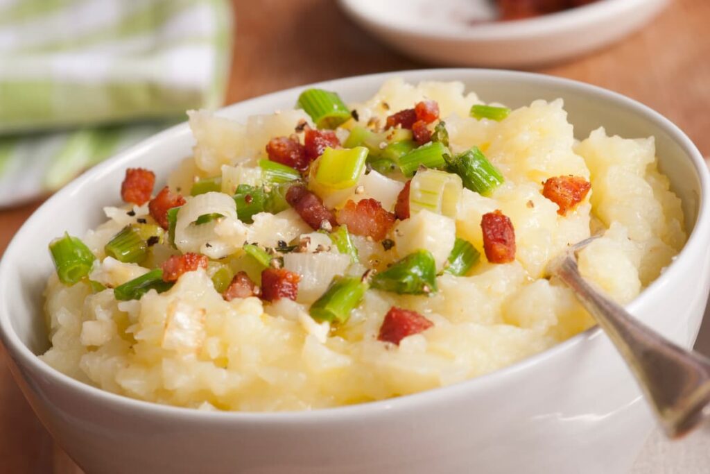 Chick Fil A Mashed Potatoes can be served in a white bowl along with the spoon. Chick Fil A mashed potato garnished with onion slices, spring onion slices, fried bacon slices.