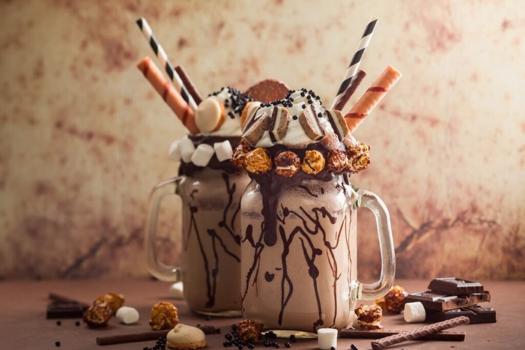 Chick Fil A Milkshakes serves in a long glass jar. There are 2 Chick fil a milkshakes which has ice cream, milkshake, chocolates, nuts and some are on the floor.