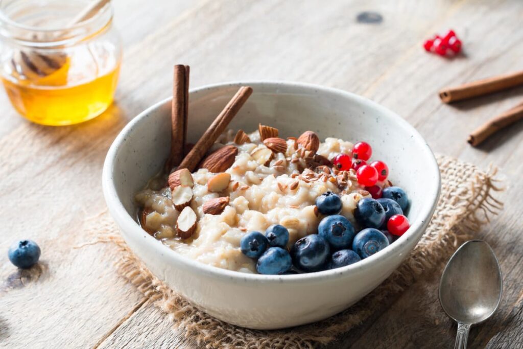 Chick Fil A Oatmeal served in a white bowl. It contains boiled oats, some almond seeds, some blue berries, red cherries, cinnamon sticks 2. Besides the Oatmeal Cup there is a honey in a glass cup, one spoon, one blueberry, 2 cinnamon sticks, and some red cherries.