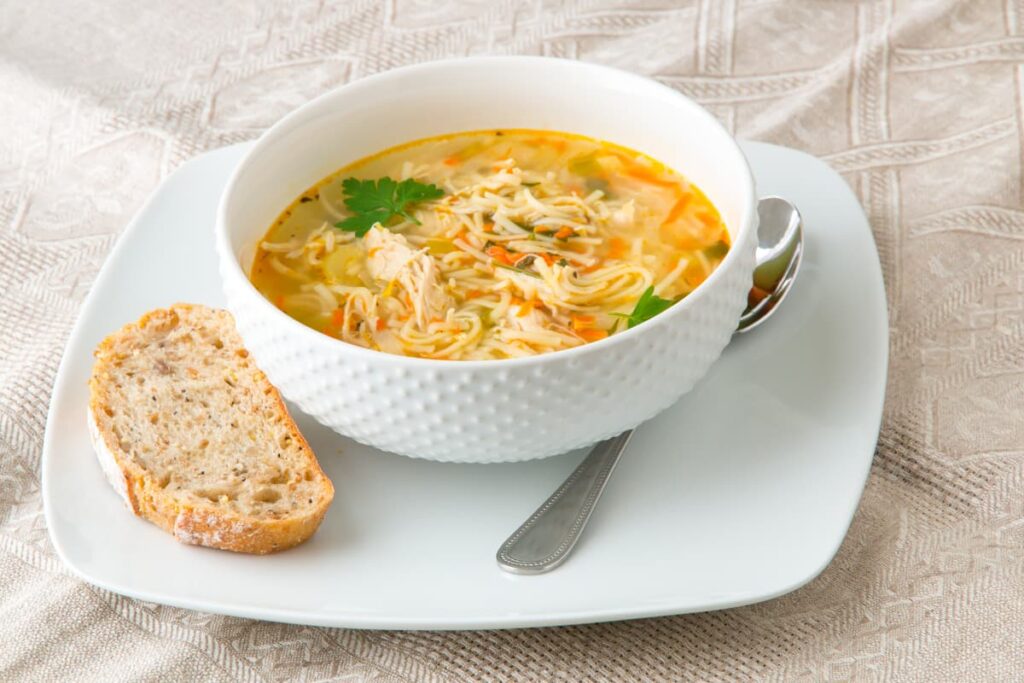 Chick Fil A Chicken Noodle Soup Serves in a white cup. This chicken noodle soup is placed in a saucer, also serves garlic bread slice and one steel spoon.