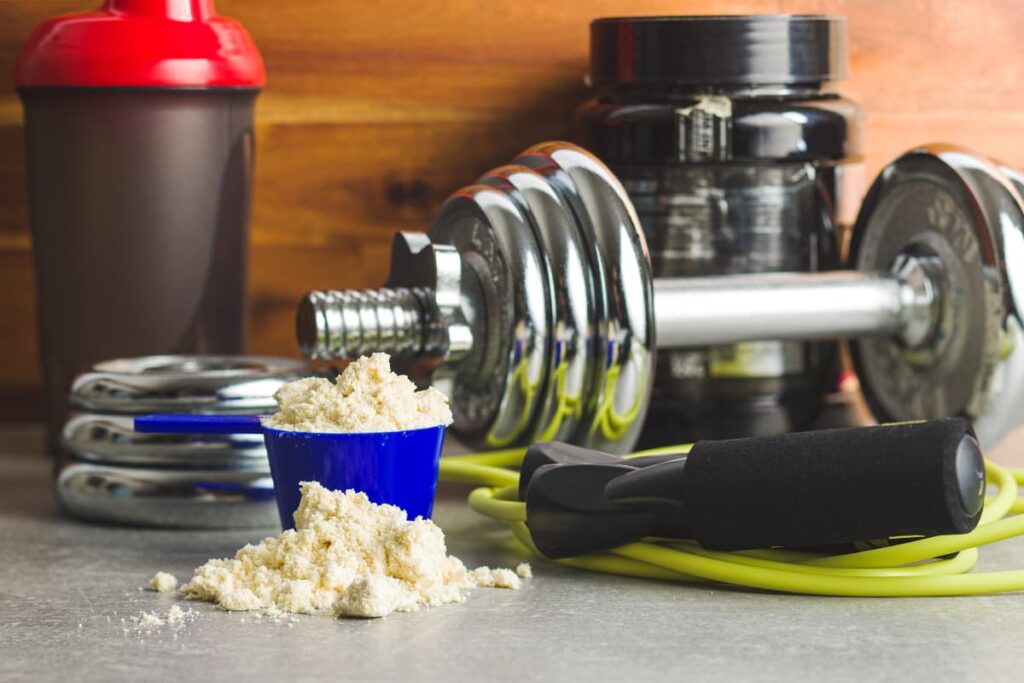 Walmart Protein Powder in a small blue cup along with blender bottle, body building equipment in a gym.