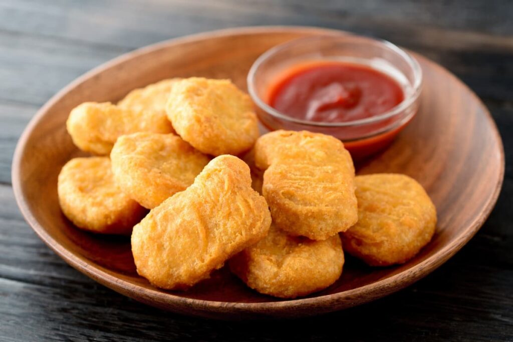 Burger King Chicken Nuggets are served in a wooden plate which is brown color. Chicken nuggets are served with the ketchup, which is red color and it is placed in small glass bowl beside the nuggets.