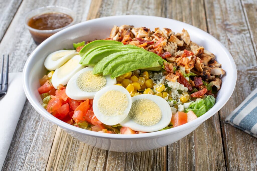 Chick Fil A Cobb Salad serves in a white plate along with the red sauce which is in a small glass bowl. Chick Fil A Salad contains boiled egg slices, tomato pieces, corn, avocado slices, fried chicken pieces, and lettuce.