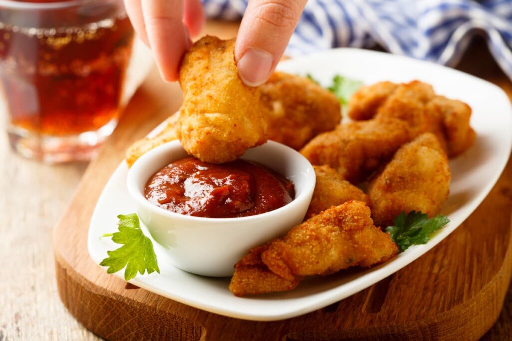 Chick Fil A Nuggets are served in a white saucer along with the red ketchup in a small white cup. A person holds chicken nugget and trying to dip into the ketchup, and the nuggets are garnished with coriander leaves. Besides serving plate you can see a red color tea which is in a glass.
