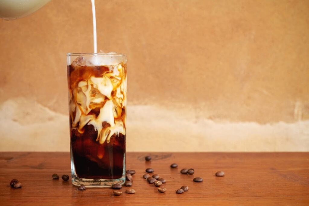 Starbucks Cold Brew serves in a glass, along with the white color straw. This Starbucks cold brew coffee includes caffeine, cream, milk, ice cubes. Beside the glass, some beans are sprinkled on the floor.