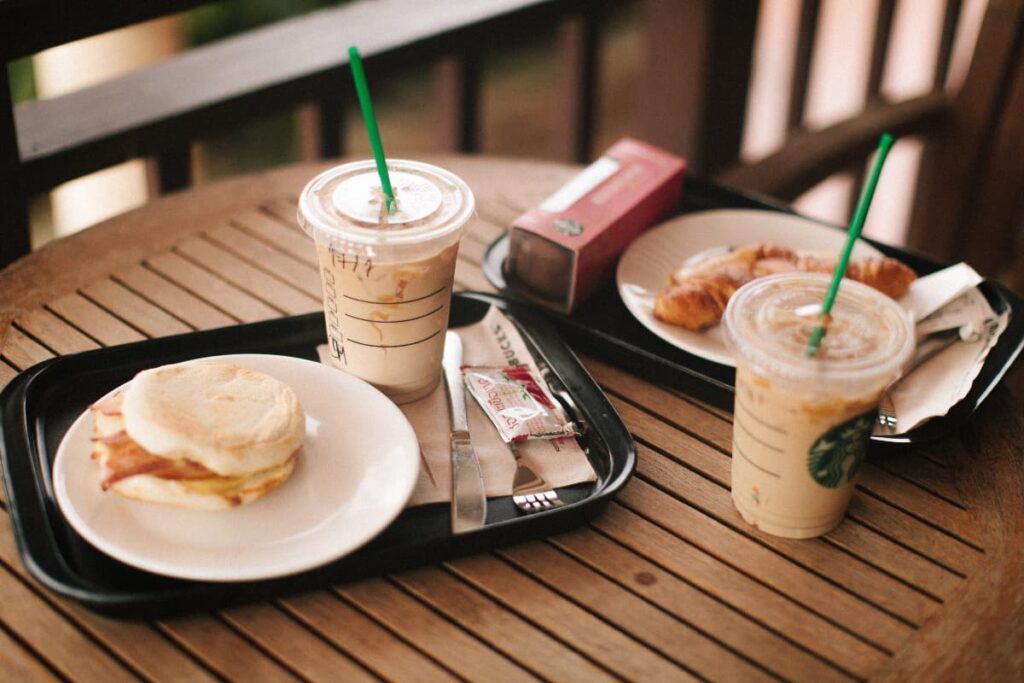 Starbucks Iced Coffee can be served in a glass along with the green straw. On a table, there are 2 trays which is having burger, puffs, ketchup, 2 forks, one knife one muffin along with the 2 starbucks iced coffee which is having starbucks logo on it.