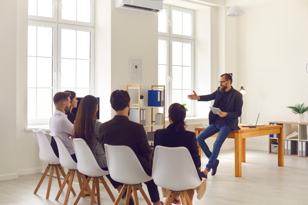 Starbucks Manager wears black blazer, black shirt and blue jeans, sat on the table by holding book on his left hand. Opposite of the starbucks manager, 5 members sat on the chairs, which is white and brown color combination.