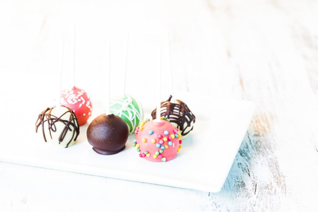 Starbucks cake pops are comes in different flavors. Starbucks cake pops are different in color which are chocolate, vanilla, straw berry, pista flavors, all are garnished with chocolate sauce, and sprinklers. All cake pops are served in a white plate.