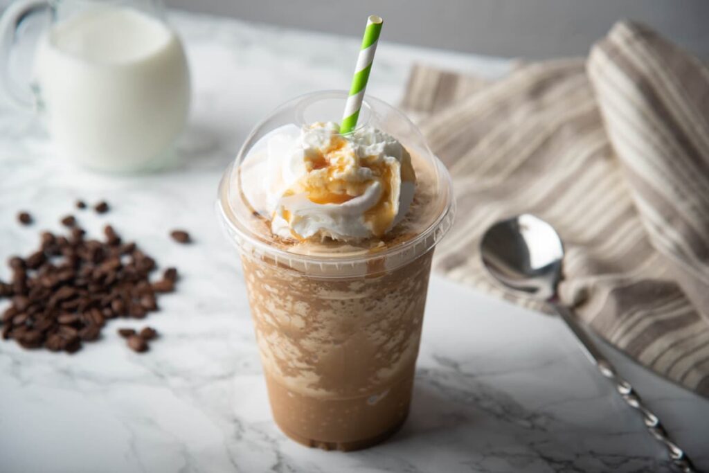 Starbucks Caramel Frappuccino serves in a glass along with the straw. Starbucks caramel frappuccino topped with white cream and garnished with the caramel. Beside the glass there are some coffee beans, one spoon, one cup of milk, one spoon and a towel.