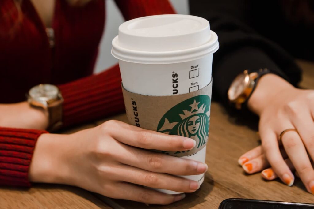 Starbucks Grande has a coffee, person holds grande coffee by her right hand who wears red t-shirt and watch on her left hand. Beside another one sitting on the table, she wears black t-shirt, watch on her right hand and a ring on her 4th finger. 