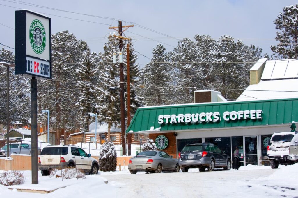 Opening a Starbucks Franchise depends on every locations. In this location, cars are parked outside the coffee shop, infront of the shop there is a starbucks logo and upside there is written Starbucks Coffee with bold letters in white color.