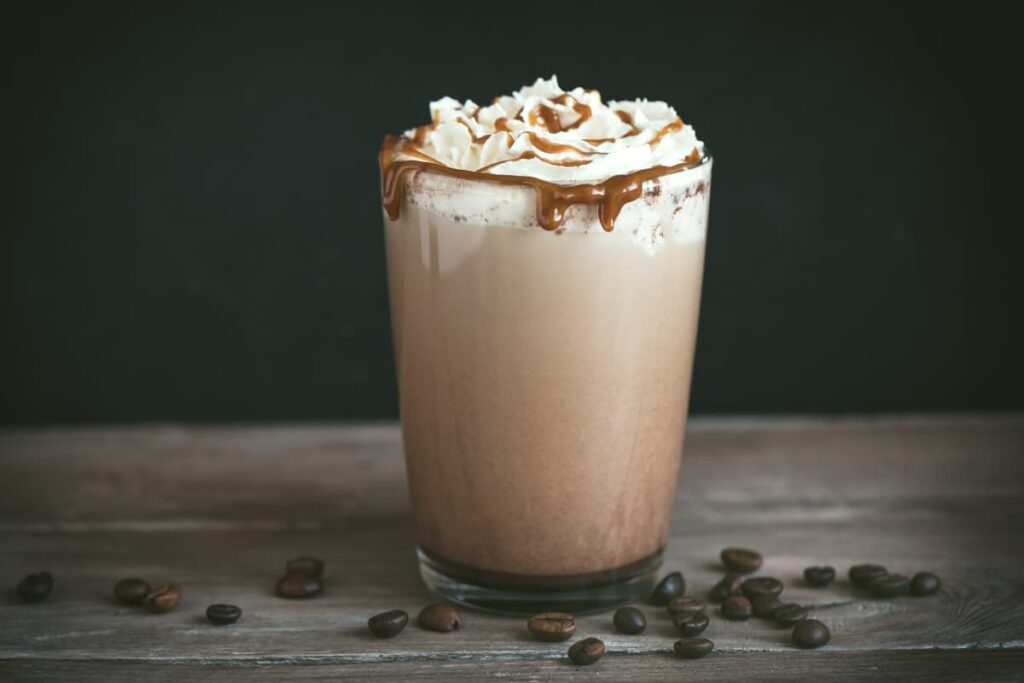 Starbucks Vanilla Bean Frappuccino can be served in a glass. Starbucks Vanilla Bean Frappuccino looks light brown color and it is garnished with whipped cream and chocolate sauce, Beside of the glass some coffee beans on the floor.