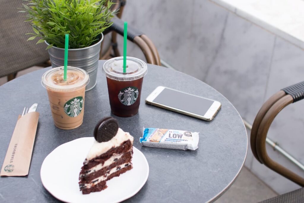 Starbucks Coffee Traveler can get 2 cup of cup(different flavors), one pastry, one chocolate along with the spoons. In a table there are 2 cups of coffee, one piece of pastry which is served on a saucer, spoons, chocolate, mobile, one artificial plant, and 2 chairs.