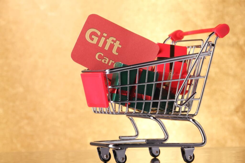 Starbucks Gift Card is placed on the shopping trolley. In shopping trolley there are some gift boxes, which are green and red color. Gift Card is mentioned in gold color at the red card and it is placed on the trolley.