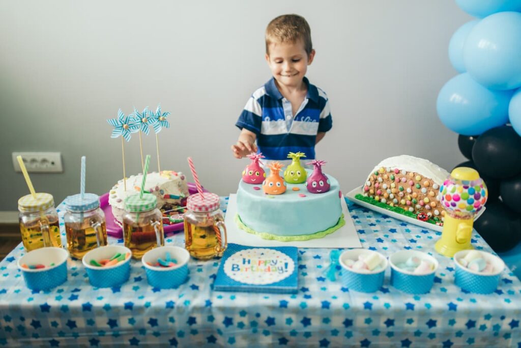 Get Free Starbucks Cakes On Your Birthday, In a table there are 3 cakes which are decorated with the gems and cream. A boy wearing blue and white T-shirt holding cake on his right side. On the table there are 4 drinks in a jars along with the 4 straws. Besides 6 blue color cups which is having marshmallows and candies. And a greeting card infront of the cake.