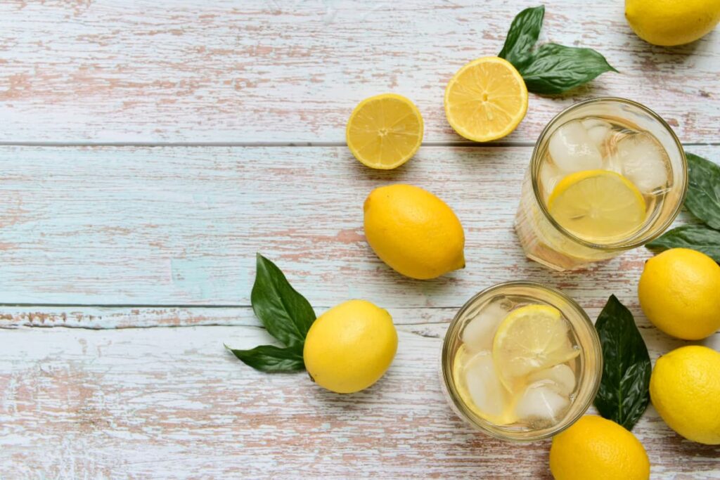 Chick Fil A Lemonade was prepared by lemons, ice cubes and sweet/salt. On a table there are some lemons, lemon slices, lemon leaves and 2 glasses of lemonade. Lemonade is garnished with lemon slices and ice cubes.