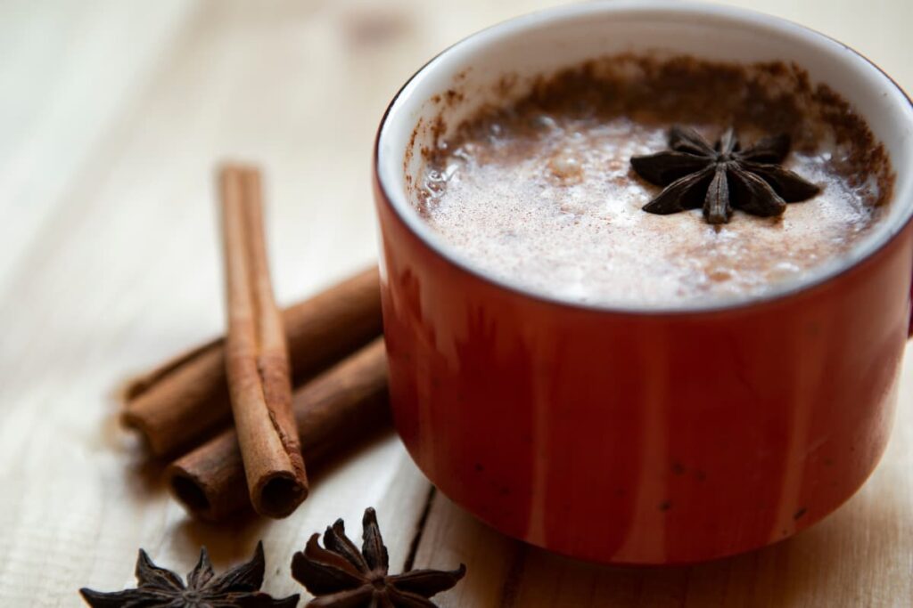 Starbucks Chai Tea Latte serves in a red color cup and it is garnished with star anise flower. Beside there are some cinnamon sticks and some anise flower on the table. 