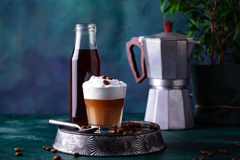 Starbucks Cold Foam serves in a glass, inside a glass half is filled with coffee on top of it cold foam and it is garnished with coffee beans. There is a coffee maker which is alluminium in color and a bottle of coffee beside the glass. Below the glass there is an ash color tray which is having coffee beans along with the spoon.