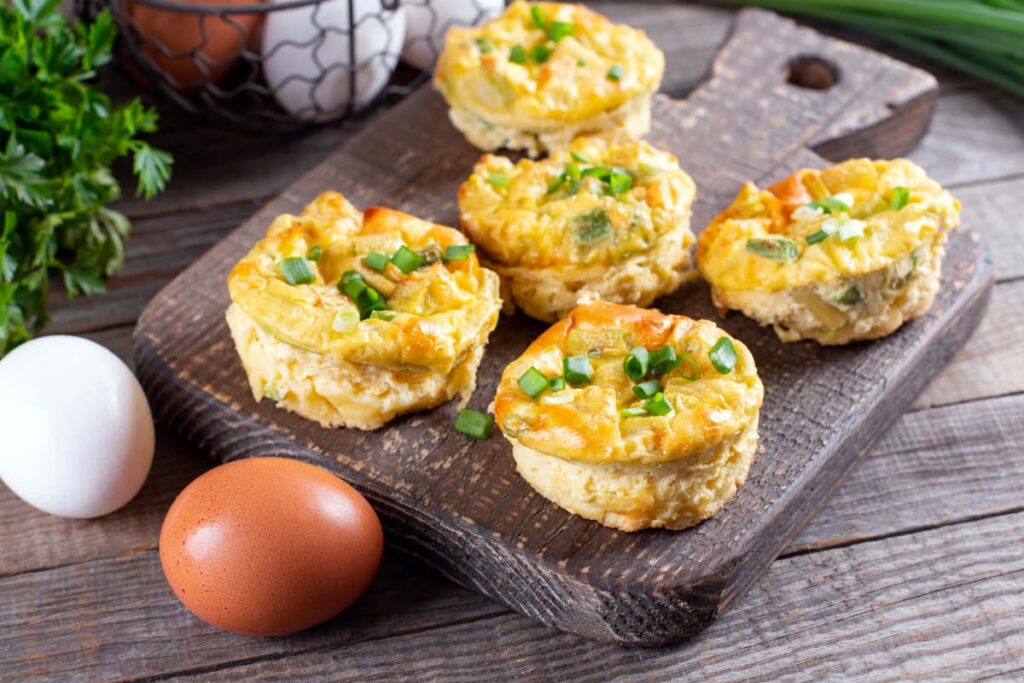 Starbucks Egg Bites serves in a brown color wooden plate. In a wooden plate 5 egg bites are served, and beside 2 eggs(one is white and another is brown color), Corainder leaves, bowl of eggs, and some grass behind it.