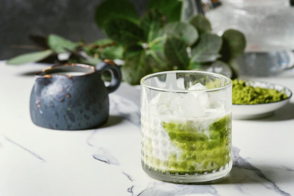 Starbucks Matcha Latte serves in a glass along with the ice cubes. Besides there is a black cup of milk, green leaves and matcha powder on the floor.