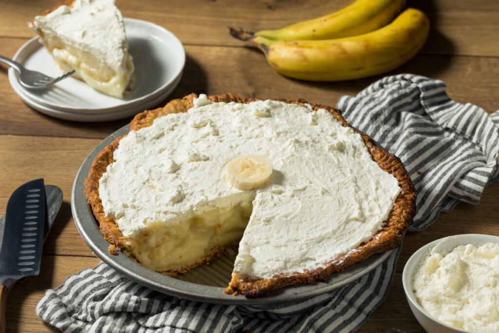 Starbucks Sweet Cream serves on top of the cake. The cake is served in a aluminium plate, beside there is a slice of cake on top sweet cream along with the fork. Beside the cake there is a white cup of sweet cream, 2 knifes and 2 bananas.