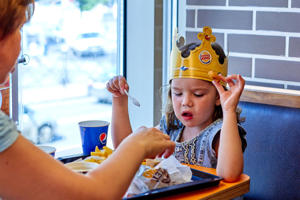 One Girl sat on the bench and eating something with the spoon. She wears Burger King Crown on her head and she wear blue colour dress. 