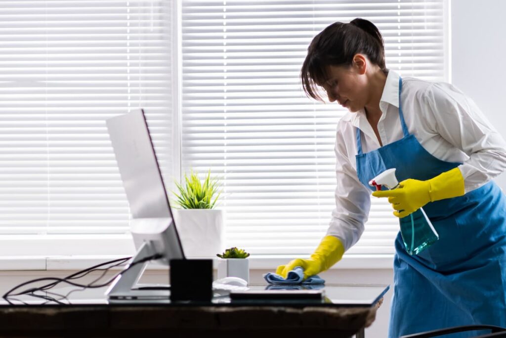 Walmart Janitor who is wearing blue apron and surgical gloves cleaning the office table , desktop, flower pots with the help of Colin.