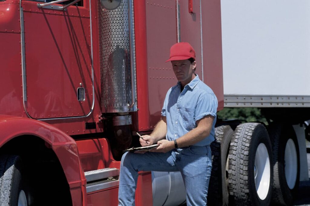 Walmart Truck Driver who wears red color cap and blue uniform writing shipment details on a paper with pen and loading boxes into truck at store.