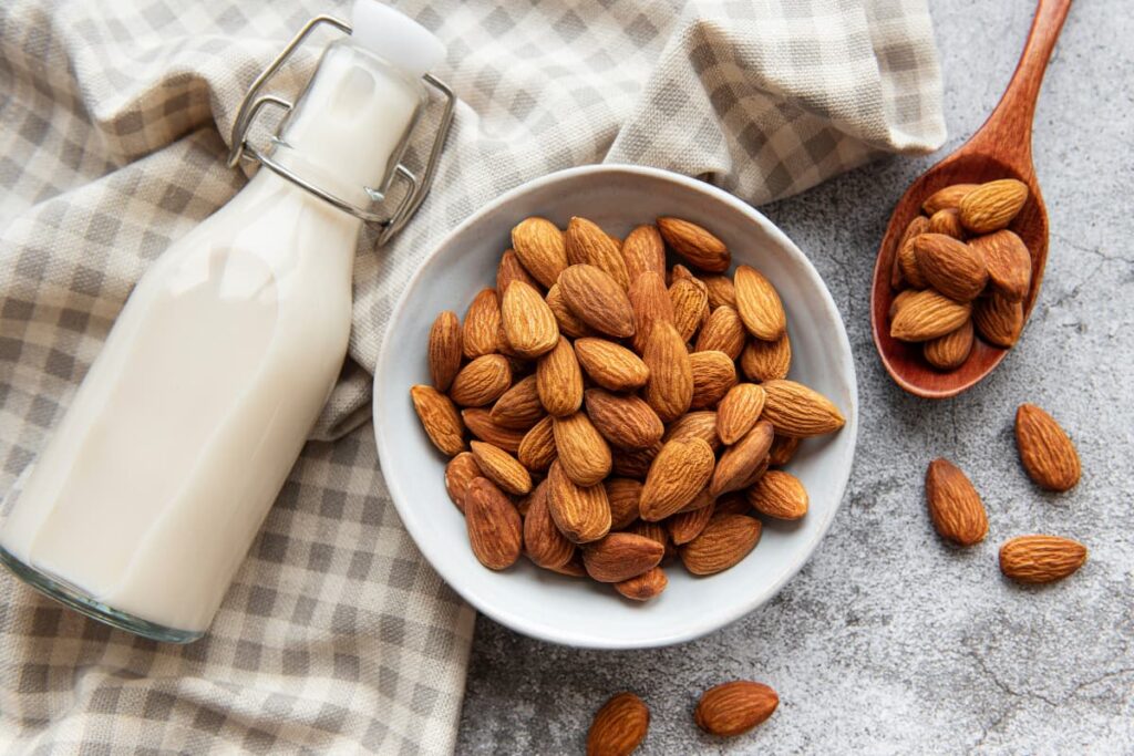 Starbucks Almond Milk serves in a glass bottle, this almond milk has been kept in a towel. On the floor some almonds are sprinkled, some are in the brown color wooden spoon and remaining almonds are kept in a white cup.