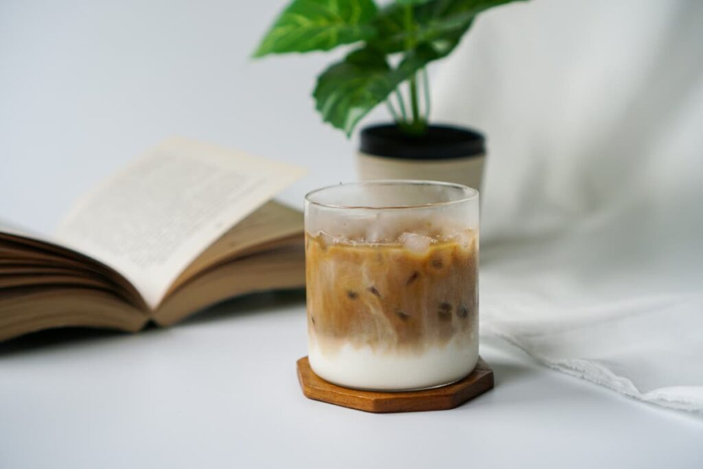 Starbucks Blonde serves in a glass along with the brown color saucer. Starbucks Blonde looks white color at the bottom and brown color on the top. Beside the blonde there is an open book and small artificial pot.