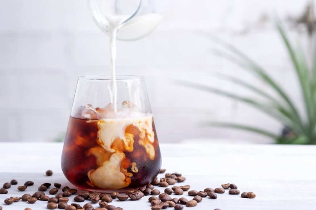 Starbucks Cold Brew coffee can be served in a large glass. Starbucks Cold Brew is a mixture of ice cubes, coffee and milk. Beside the glass there are some coffee beans on the floor, another glass of milk was pouring into the mixture.