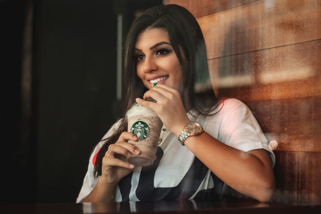 Starbucks Handcrafted Drink can be served in a glass along with the straw. A girl sat on the chair and drinking handcrafted drink, she wears white and black color combination t-shirt, and she holds drink by her right hand and she wear watch by her left hand.