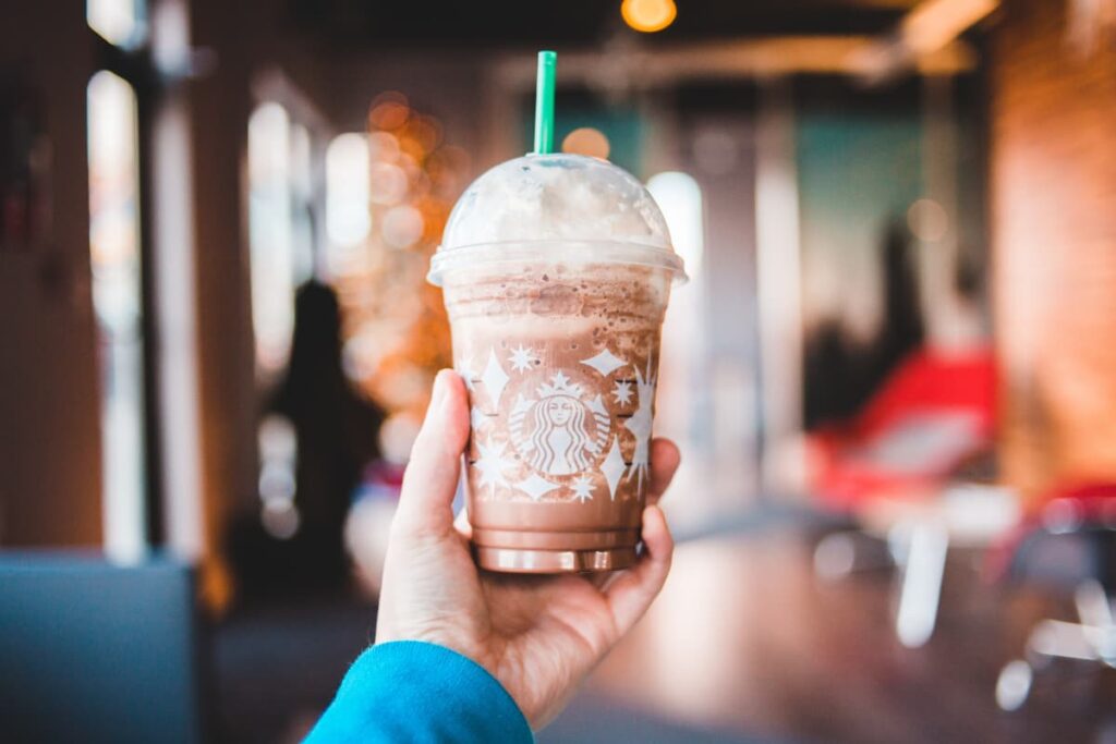 Starbucks Long Shot serves in a Starbucks glass along with the straw. A person who wears blue shirt holds Starbucks Long Shot with his/her left hand.