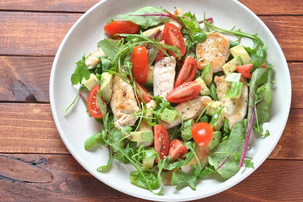 Walmart Chicken served with tomato slices, pepper, Basil leaves, Coriander leaves and avocado pieces in a white serving plate on a wooden table.