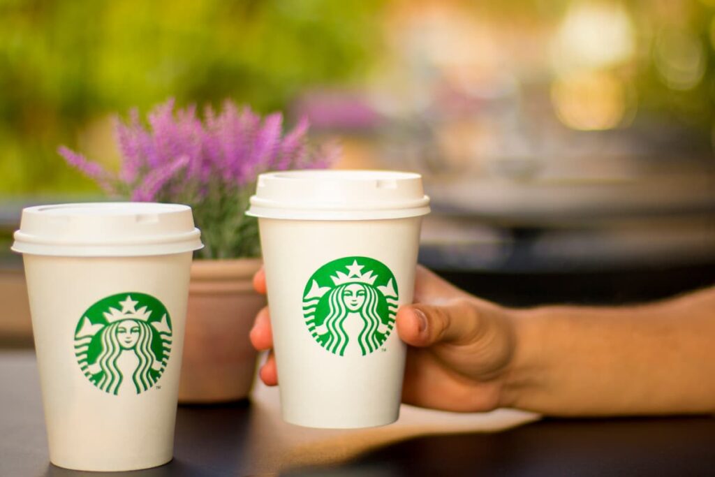 Starbucks Logo represents a mermaid which is green and white color combination. This Starbucks logo can fit into small, large, medium cups. A person holds starbucks cup with his right hand on the table, beside there is another starbucks cup, backside one flower vase is there on the table.