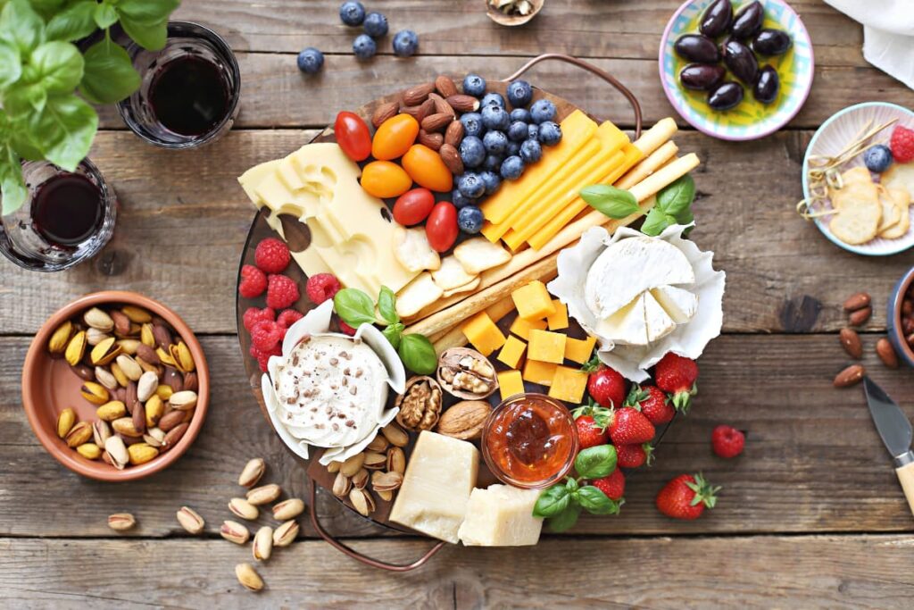 Platter at Walmart store consists of bowl of dry fruits almonds, pistachios, one bowl of java plums, one bowl of pine apple slices, one big bowl of strawberries, cane berries, rasp berries, cheese slices, wall nuts, mango cubes, paneer cubes and two glasses of wine on a wooden table. 