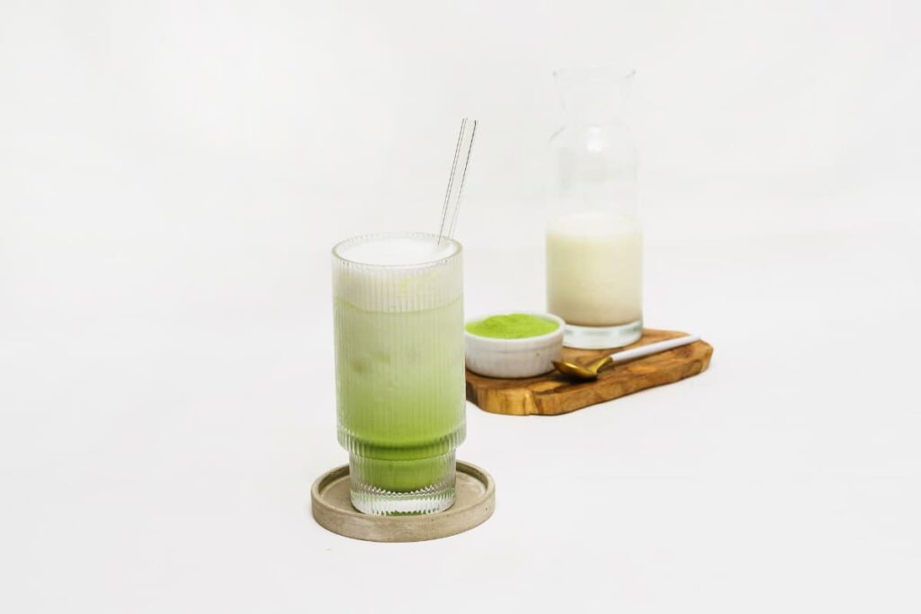 Starbucks macha serves in a glass along with the straw. This starbucks macha looks green in color at the bottom and at the top it is white in color. Beside this there is a wooden plate which is having matcha powder in a small white cup, milk in a glass along with the spoon.