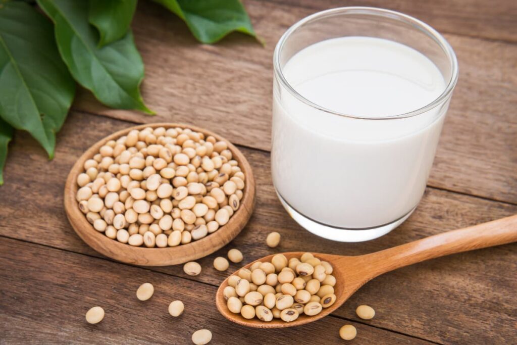 Starbucks Soy Milk looks white in color, and it is served in a glass. On atable there are plate of soya beans and spoon of soya beans and some are on the floor, beside there are some leaves on the floor.